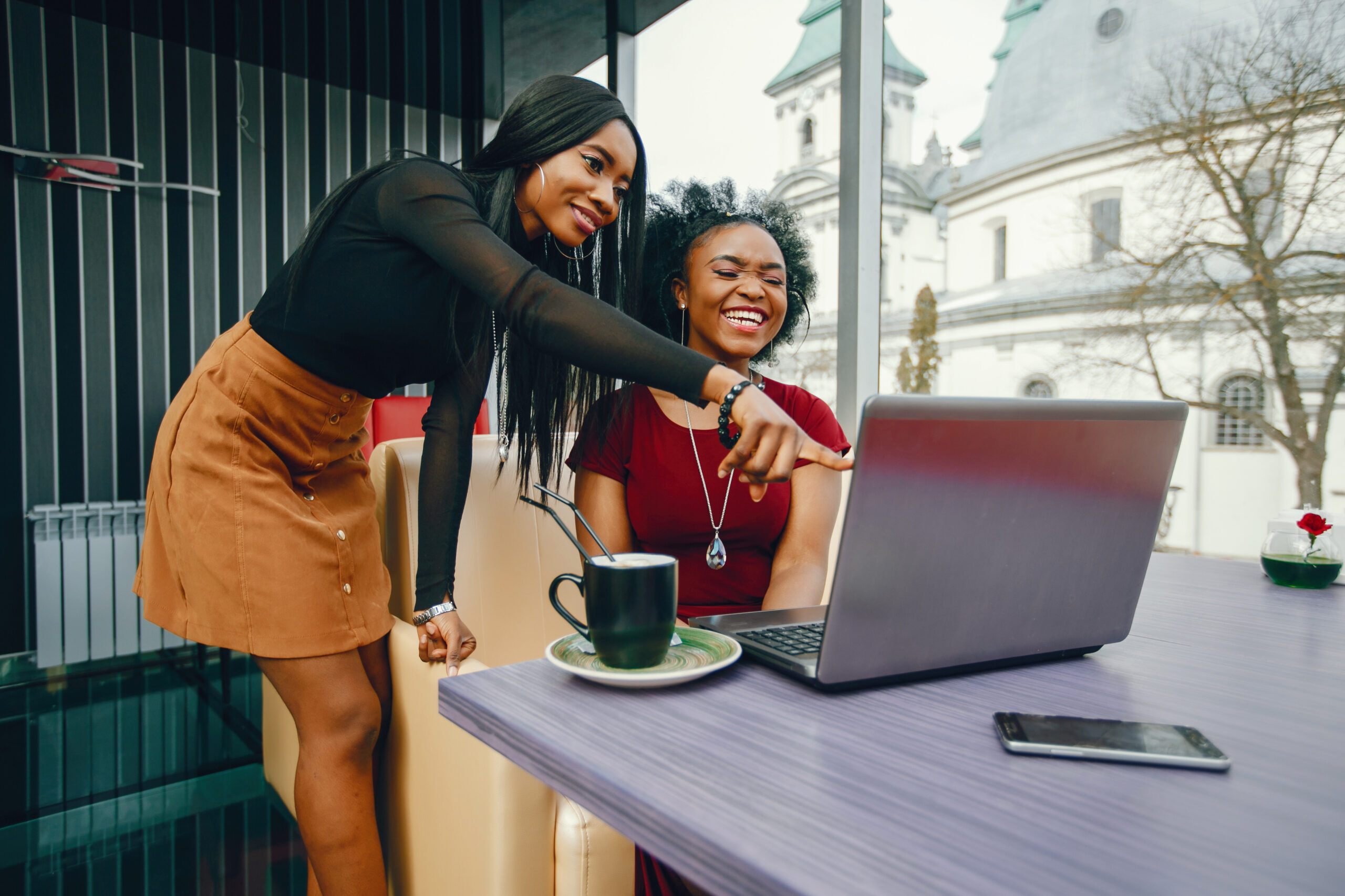 two beautiful and stylish young, dark girls sitting in a restaurant at the table, chatting and using a laptop and drinking a coffee